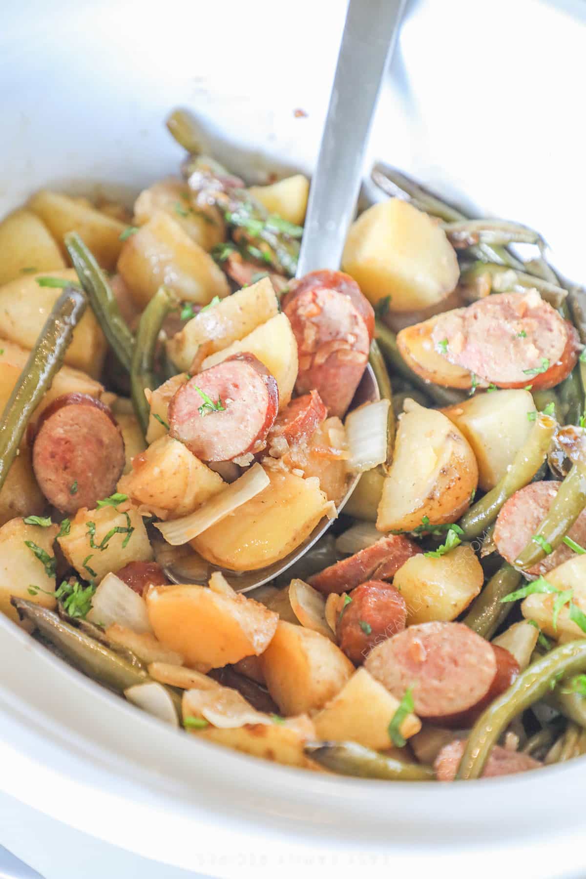 A white Crock Pot full of sausage and potatoes with a silver serving spoon.