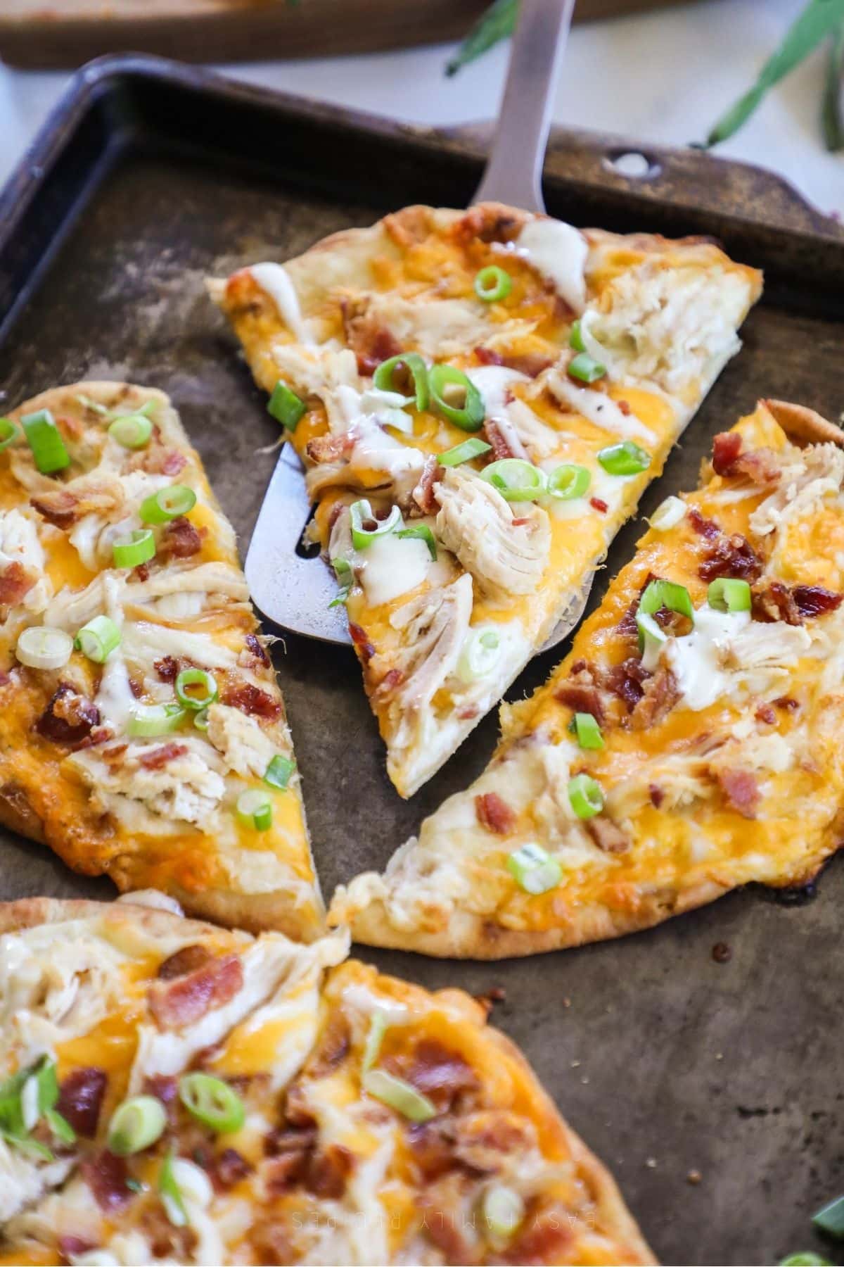 chicken bacon ranch flatbread on a baking sheet, cut, with a slice being lifted with a spatula