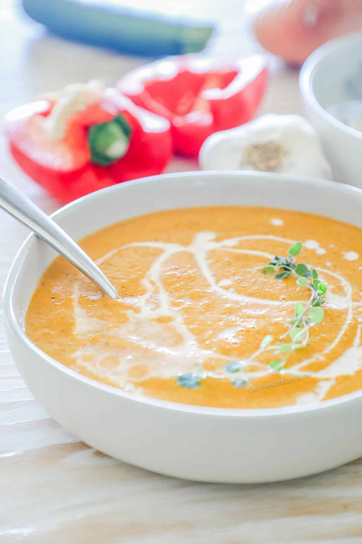 Roasted veggie soup in a white bowl with a spoon. Red bell peppers are in the background.