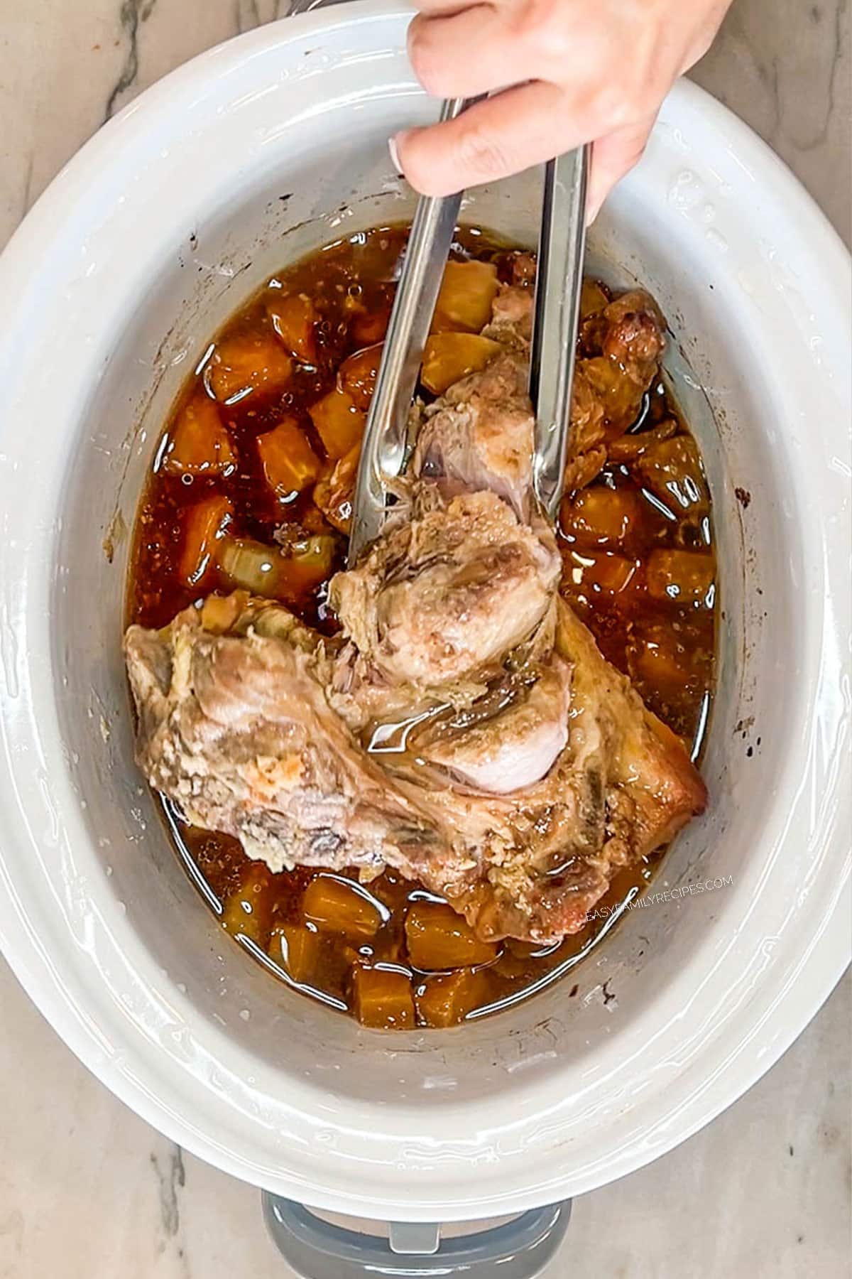 Hand using tongs to remove the cooked pork shoulder from the crockpot.