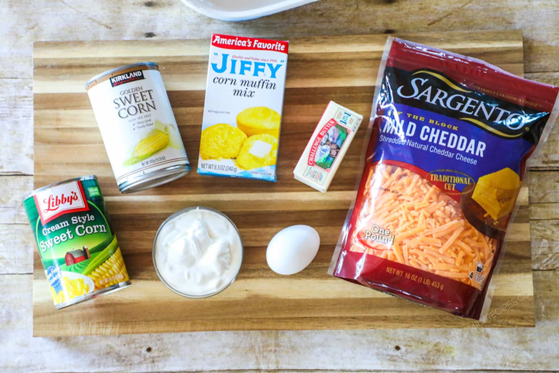 Ingredients for making Jiffy Corn Casserole including Jiffy cornbread mix, cream corn, canned corn, cheddar cheese, sour cream, eggs, and butter.