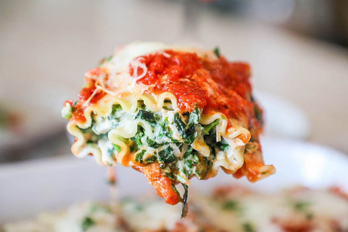 Side view of a spinach chicken lasagna roll up hovering over the rest of them in a baking dish.
