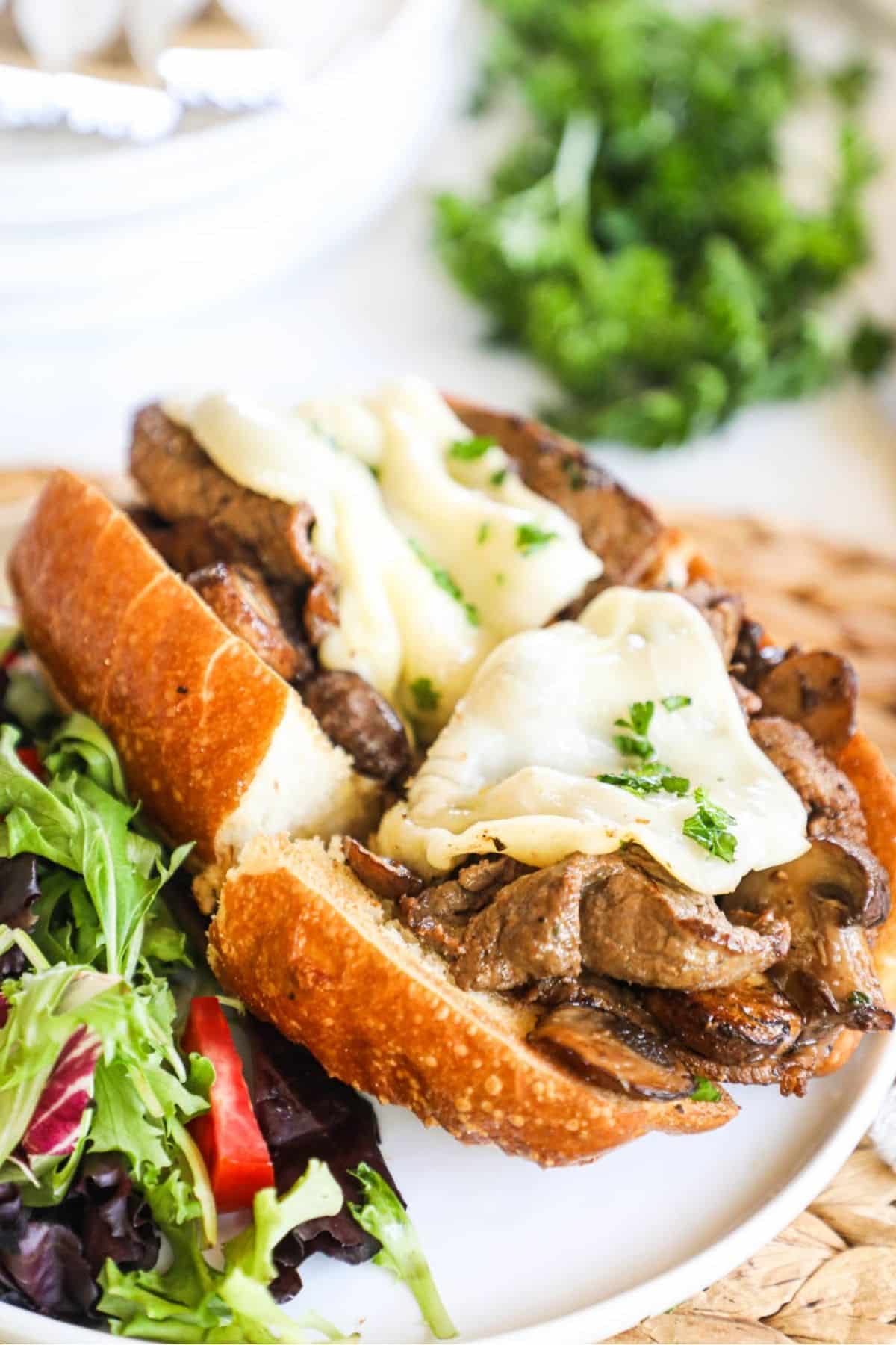 steak and mushroom sandwich topped with cheese on a plate with salad