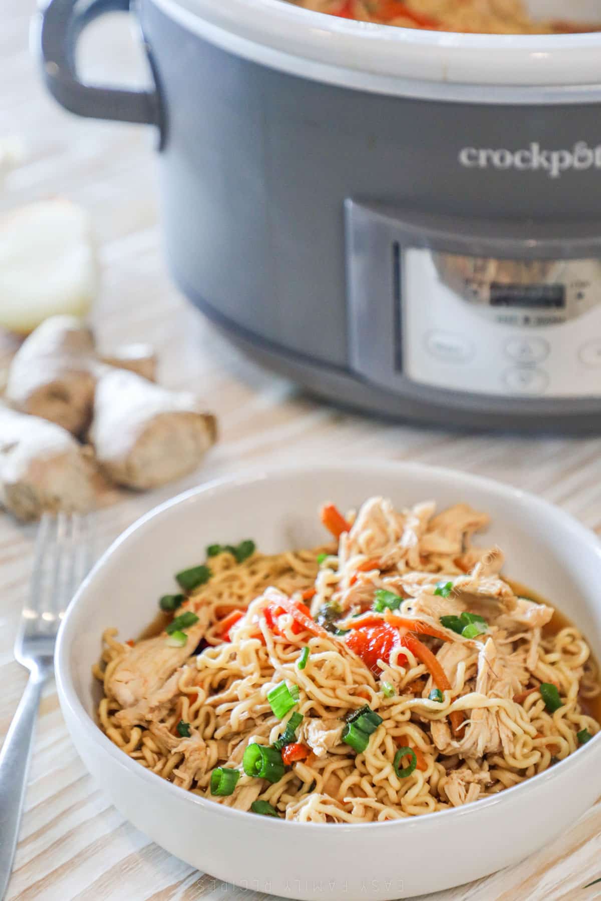 A serving of Crockpot Chicken Ramen in a white bowl with garlic and a gray Crockpot in the background.