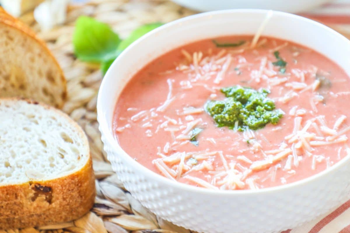 white bowl of tomato soup topped with cheese and a dollop of pesto, with a slice of fresh bread on the side