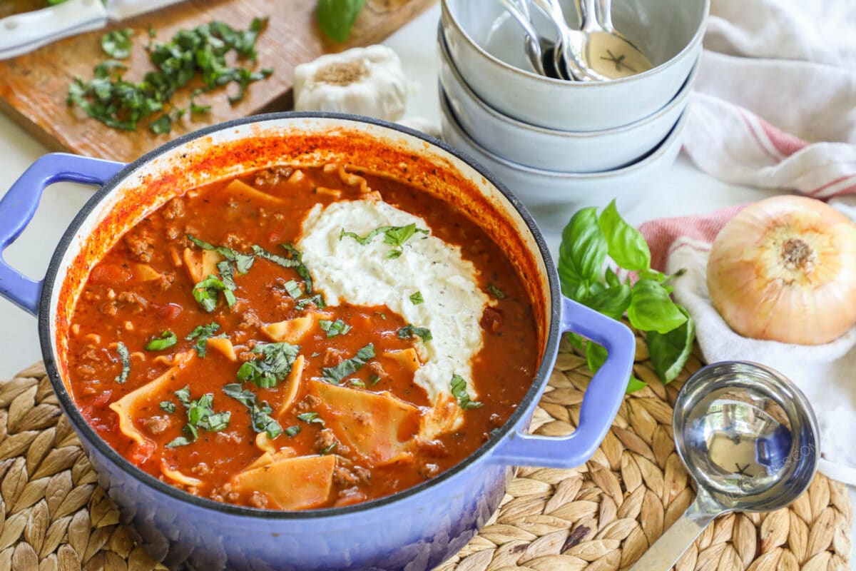 Overhead view of Lasagna Soup in a blue dutch oven on a table with bowls and an onion nearby