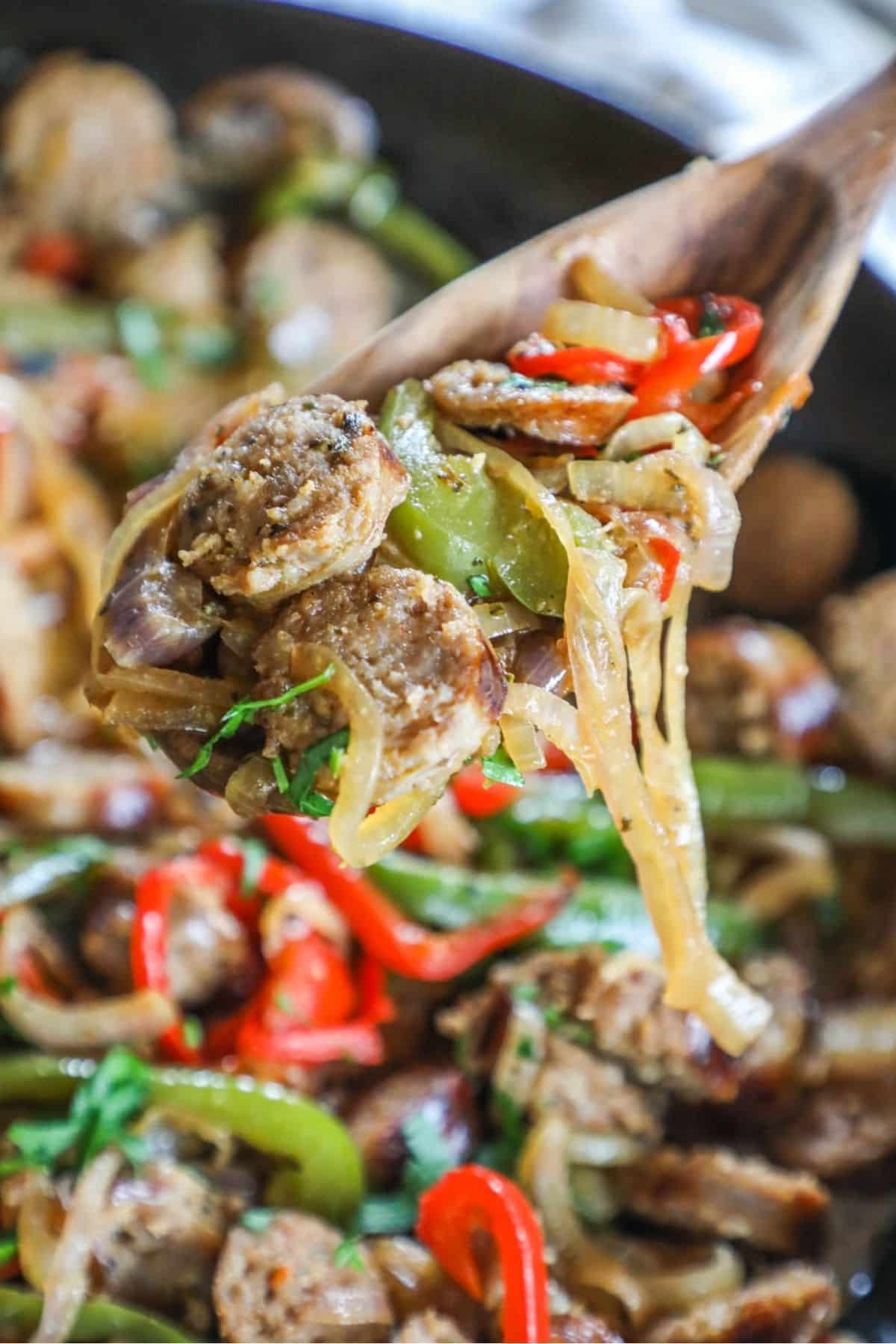 wooden spoon holding a scoop of sausage and peppers and onions lifted from a skillet