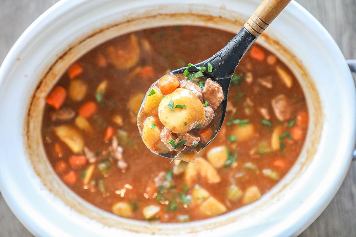 slow cooker beef stew in white crockpot with wooden spoon lifting soup out