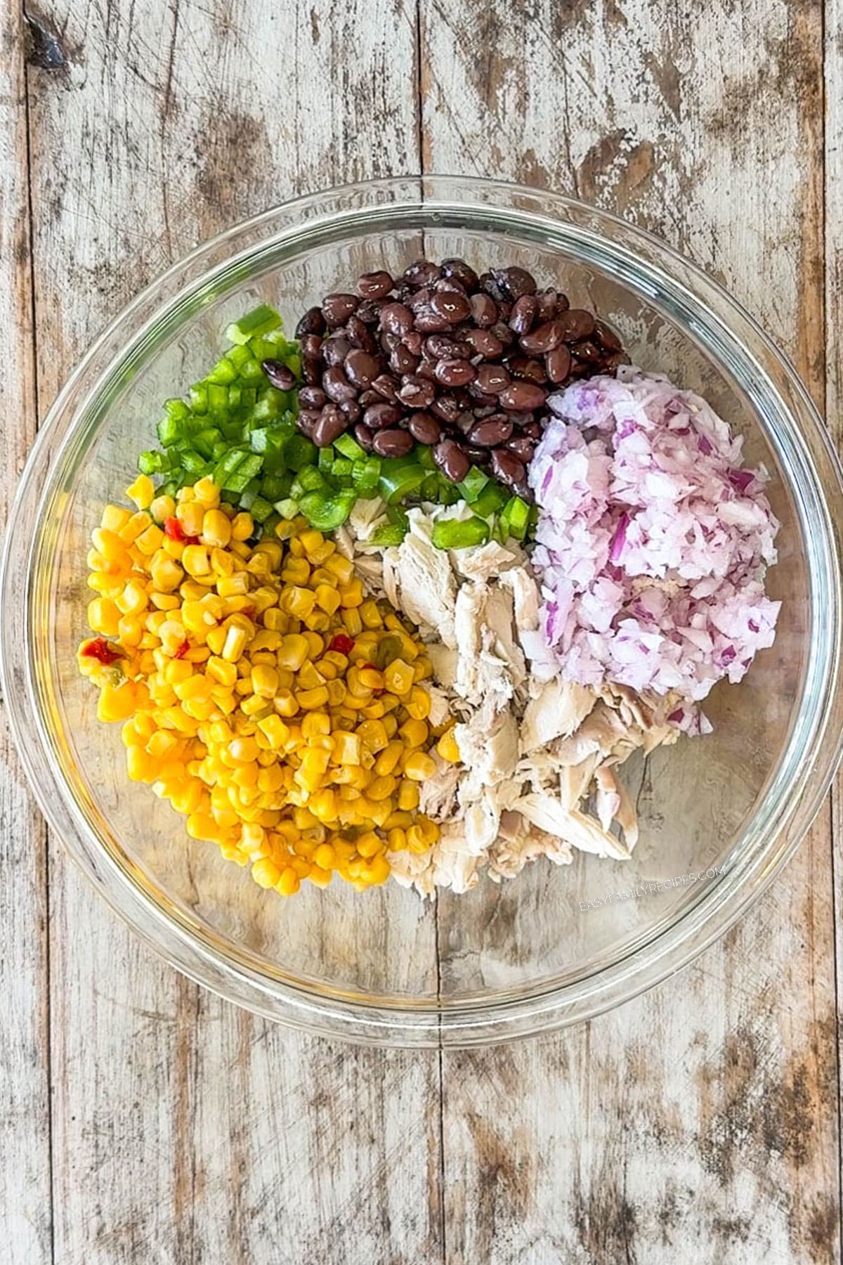ingredients for mexican chicken salad in glass bowl, before being mixed together. includes shredded chicken, black beans, corn, green pepper, red onion, mayo, sour cream, lime juice and taco seasoning
