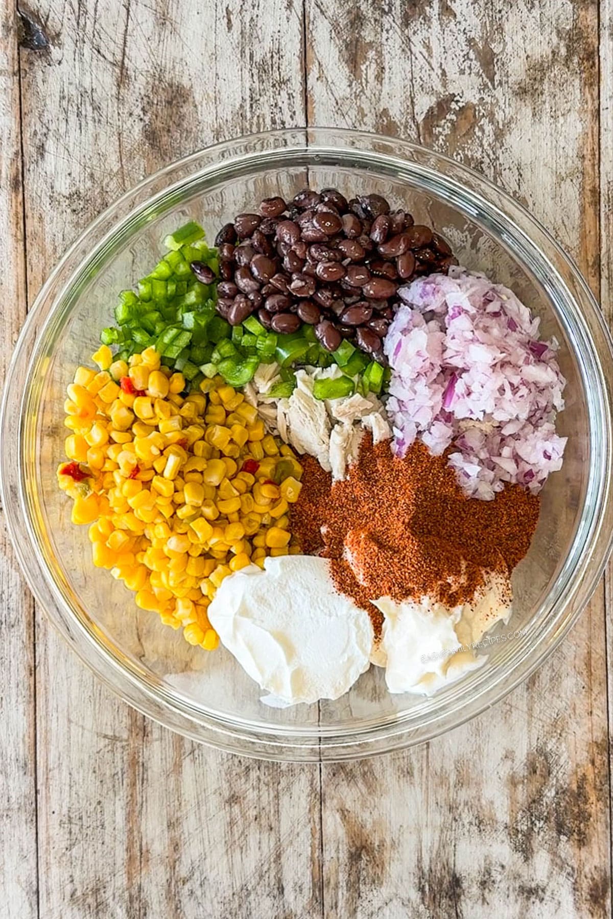 ingredients for mexican chicken salad in glass bowl, before being mixed together. includes shredded chicken, black beans, corn, green pepper, red onion, mayo, sour cream, lime juice and taco seasoning