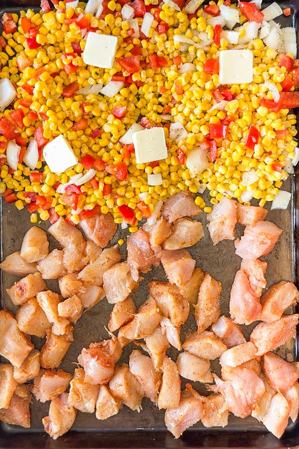Corn mixed with onions and red bell peppers with butter on top next to chicken on a sheet pan.