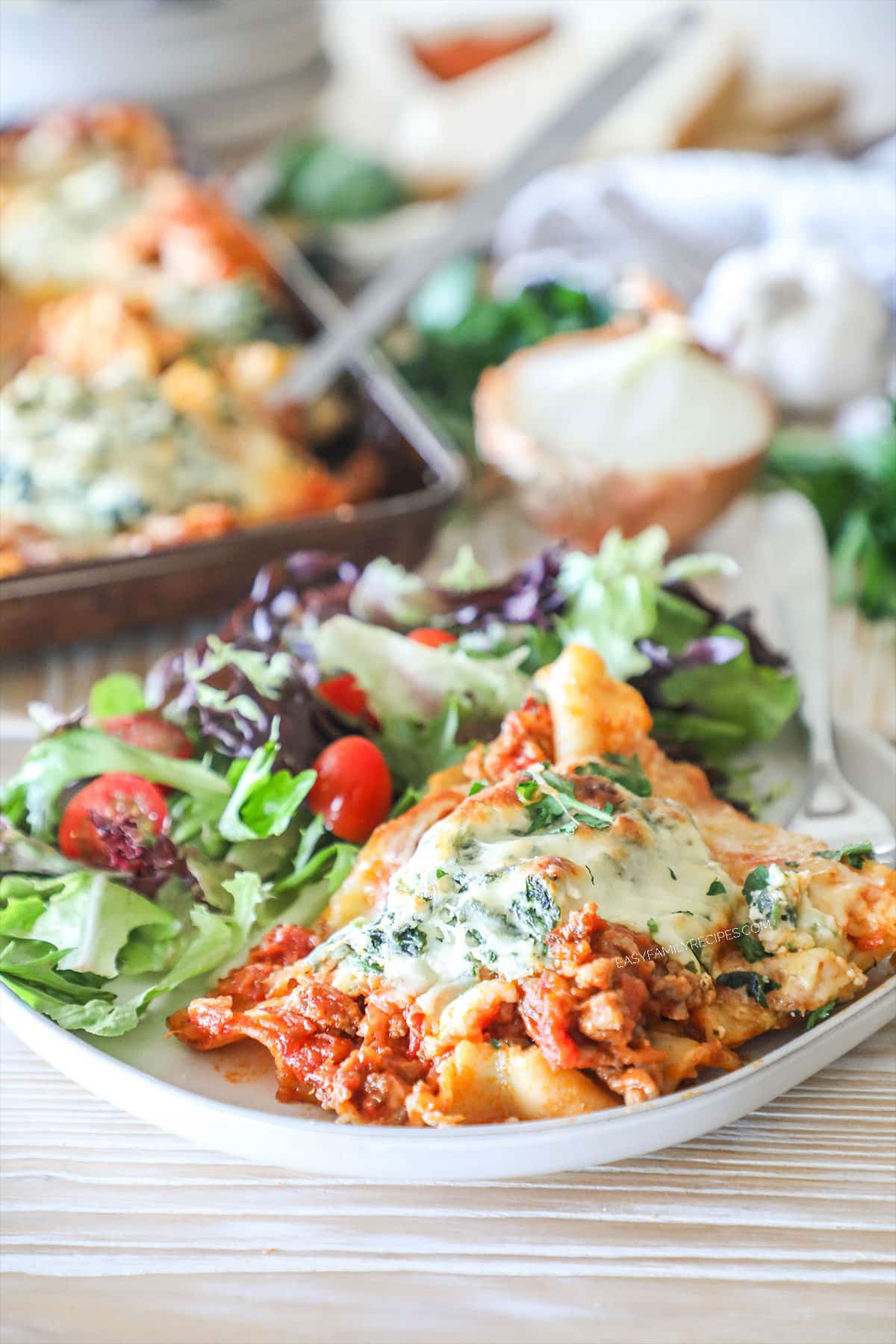 Individual slice of sheet pan lasagna on white dish with side salad. Sheet pan with lasagna in background. On wooden background with onion, garlic and fresh herbs.