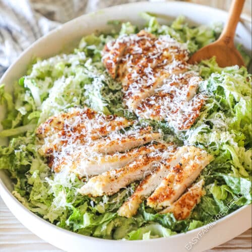 parmesan chicken breasts sliced and placed over green goddess salad in a large white bowl with two wooden spoons