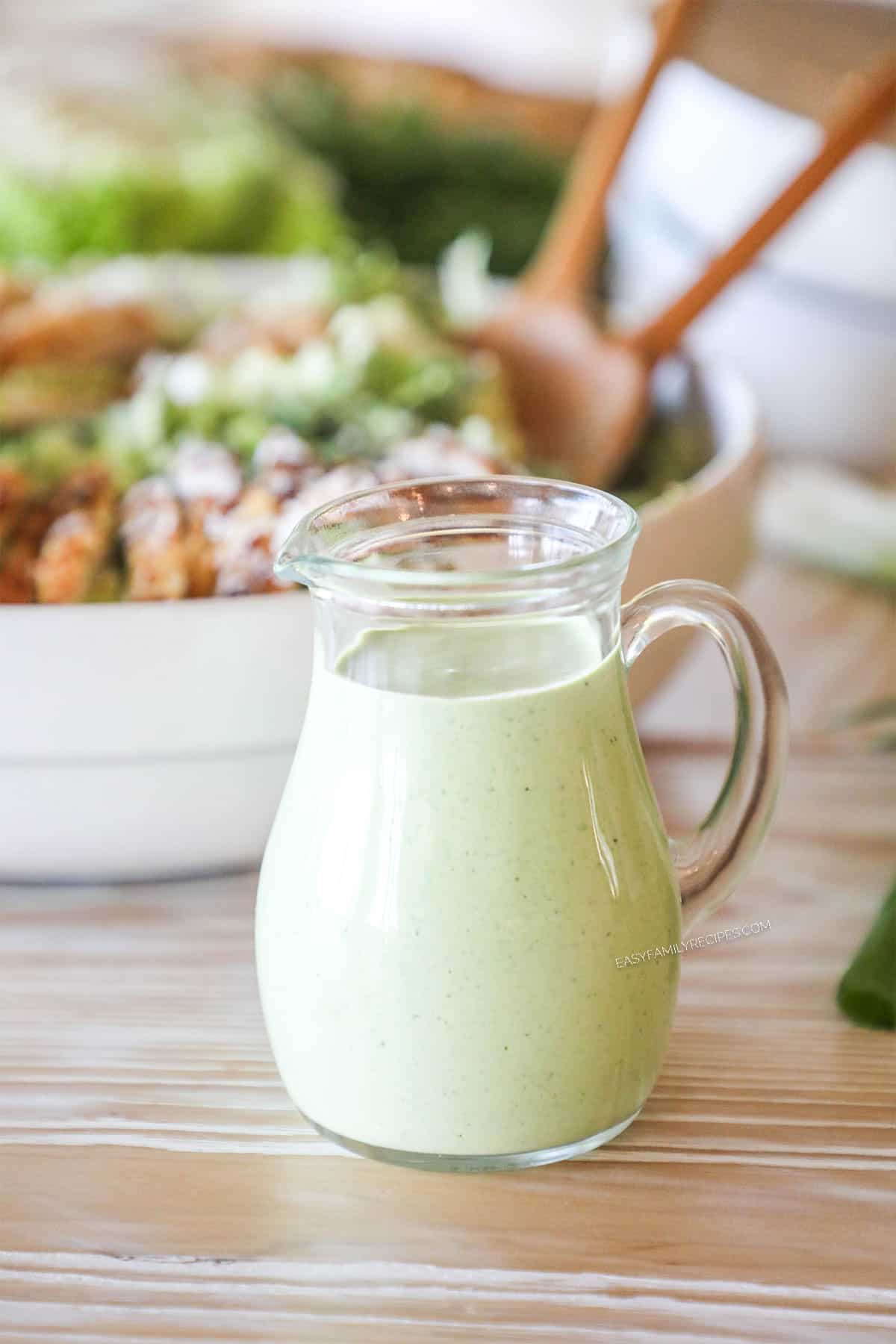 small pitcher of green goddess dressing in front of a large white bowl with completed salad and chicken breasts on top- two wooden spoons in salad