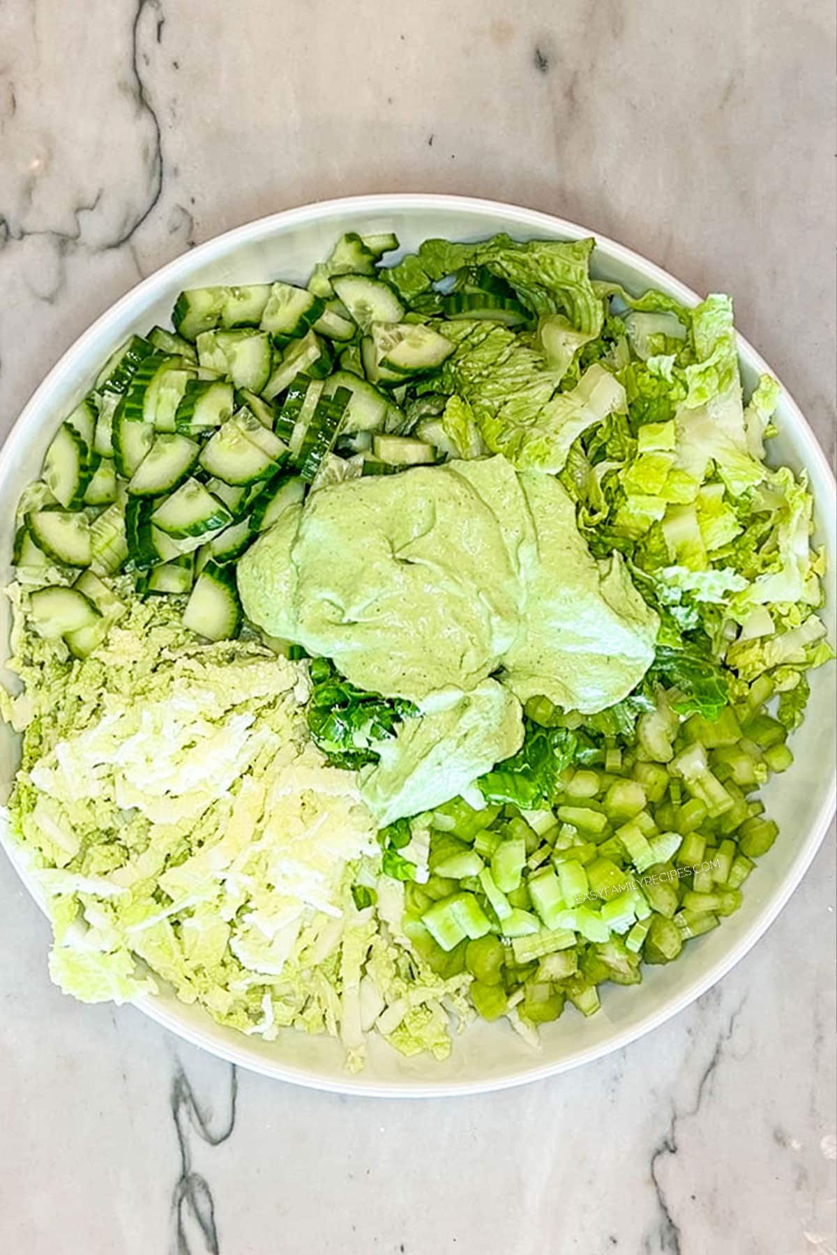 greens, veggies and dressing for green goddess salad before being mixed together- in a large white bowl with a white marble background