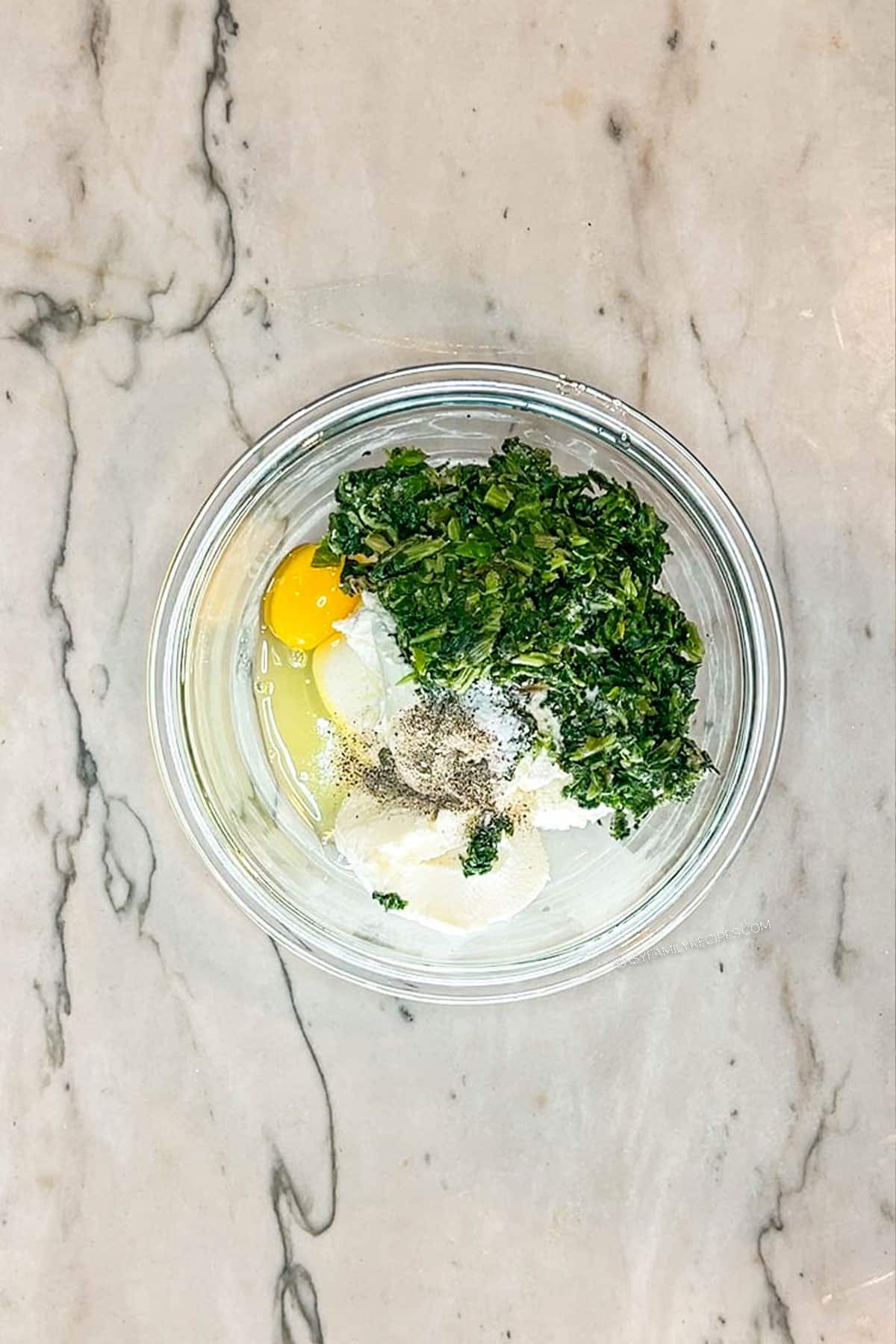 Glass bowl containing ricotta, frozen spinach, egg and ricotta. On a marble background.
