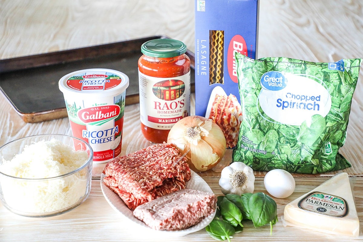 Ingredients needed to make Sheet Pan Lasagna including ground beef, Italian sausage, Marinara, noodles, ricotta, Mozzarella, onion, garlic, basil, frozen spinach and egg. On a wooden background.