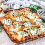Sheet pan lasagna on wooden background, surrounded by metal spatula, garlic head, basil and onion