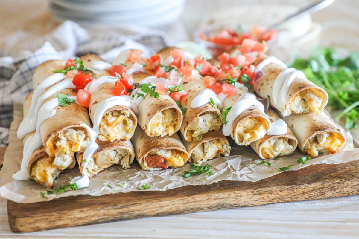 Close up of Baked Chicken Taquitos with toppings including pico and sour cream- on foil and a wooden board- fresh herbs around