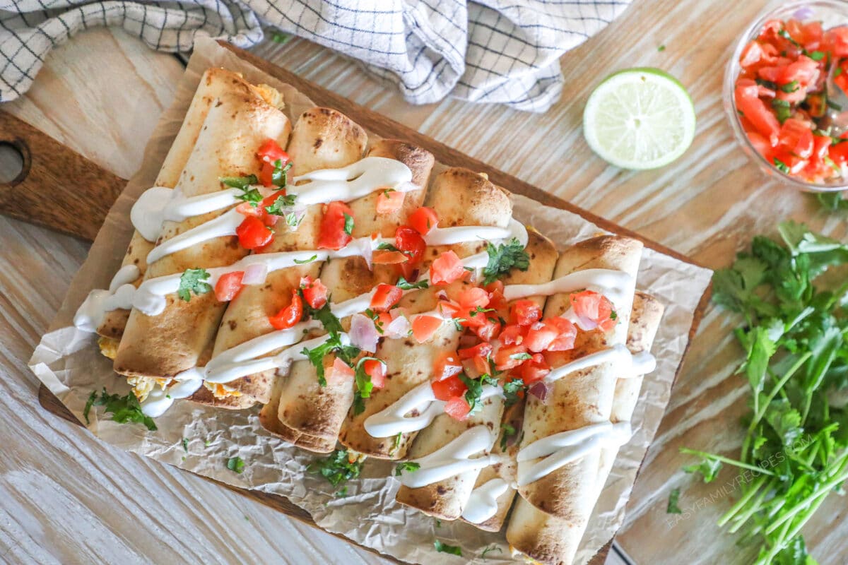 Close up of Baked Chicken Taquitos with toppings including pico and sour cream- on foil and a wooden board. On a wooden background with fresh herbs and ingredients around