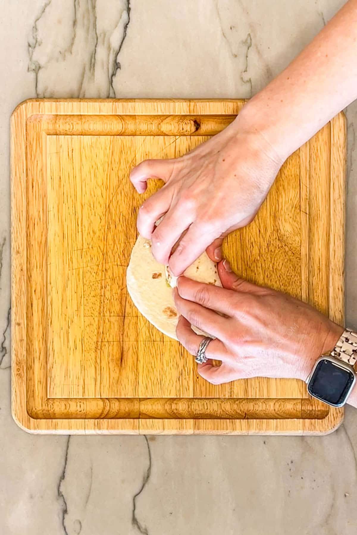 Hands folding a filled tortilla for Baked Chicken Taquitos. On a wooden board with a marble background