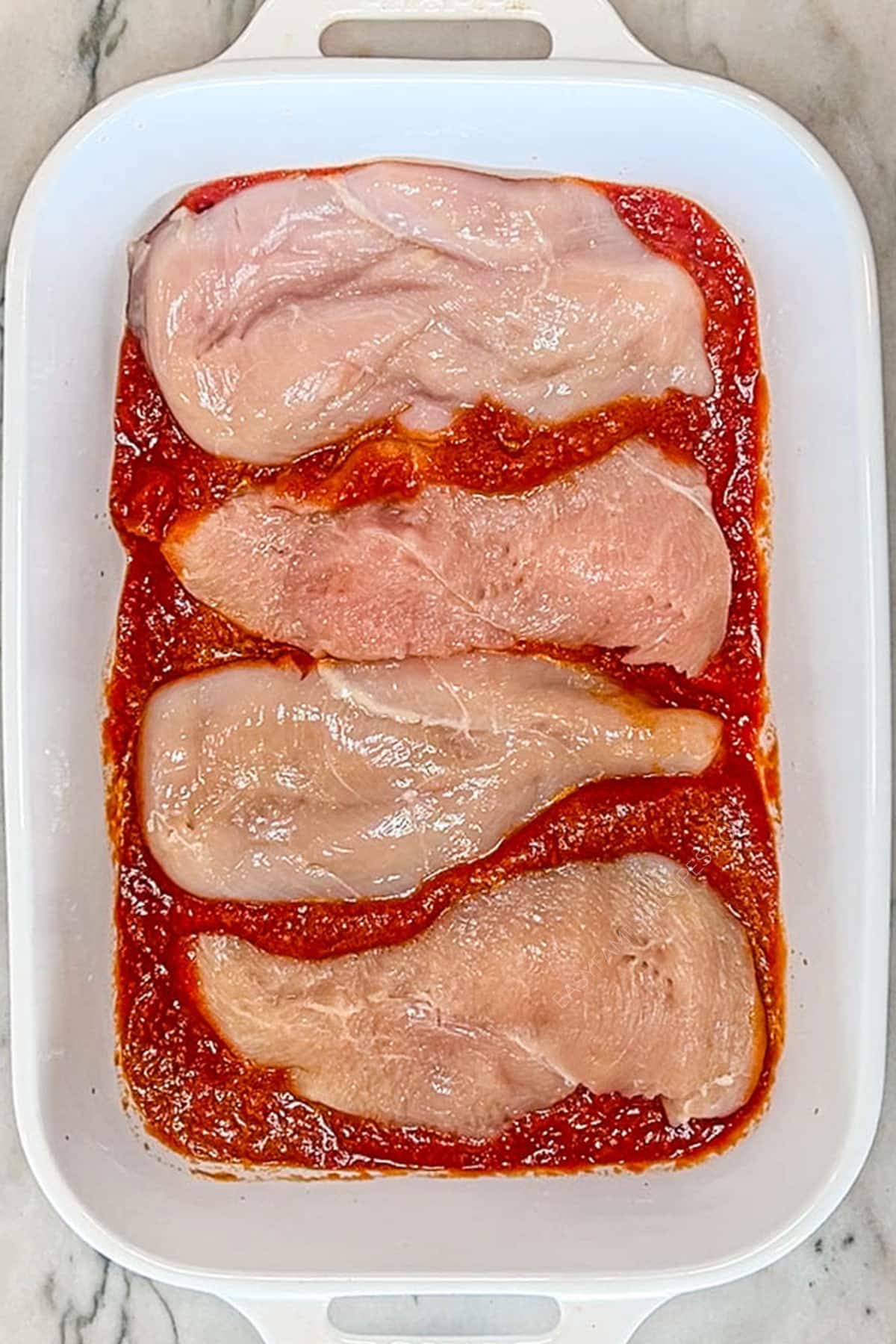 Four chicken breasts in tomato sauce in a white baking dish.