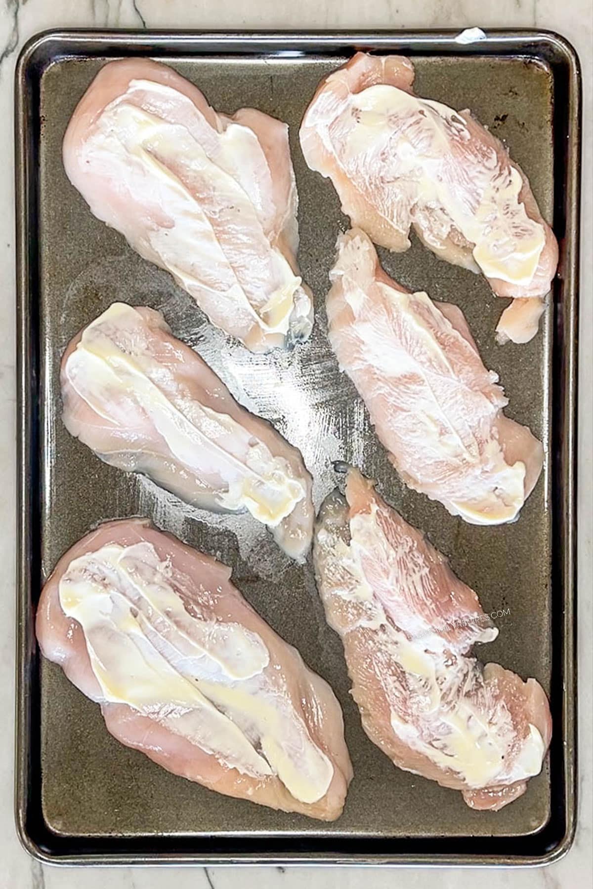 Pieces of chicken breast on a sheet pan with mayonnaise spread on top.
