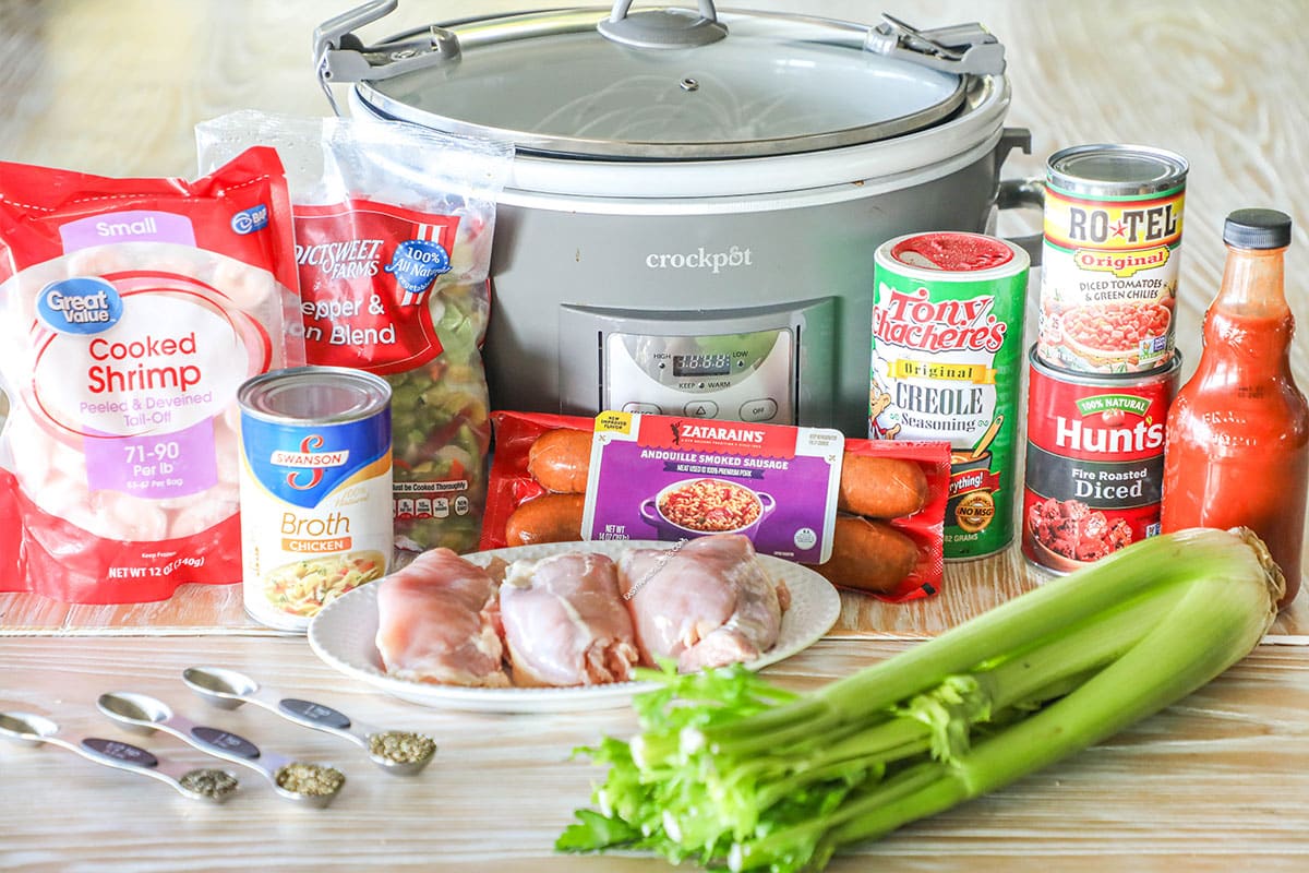 Ingredients needed to make slow cooker jambalaya- with grey slow cooker- on a wooden backdrop