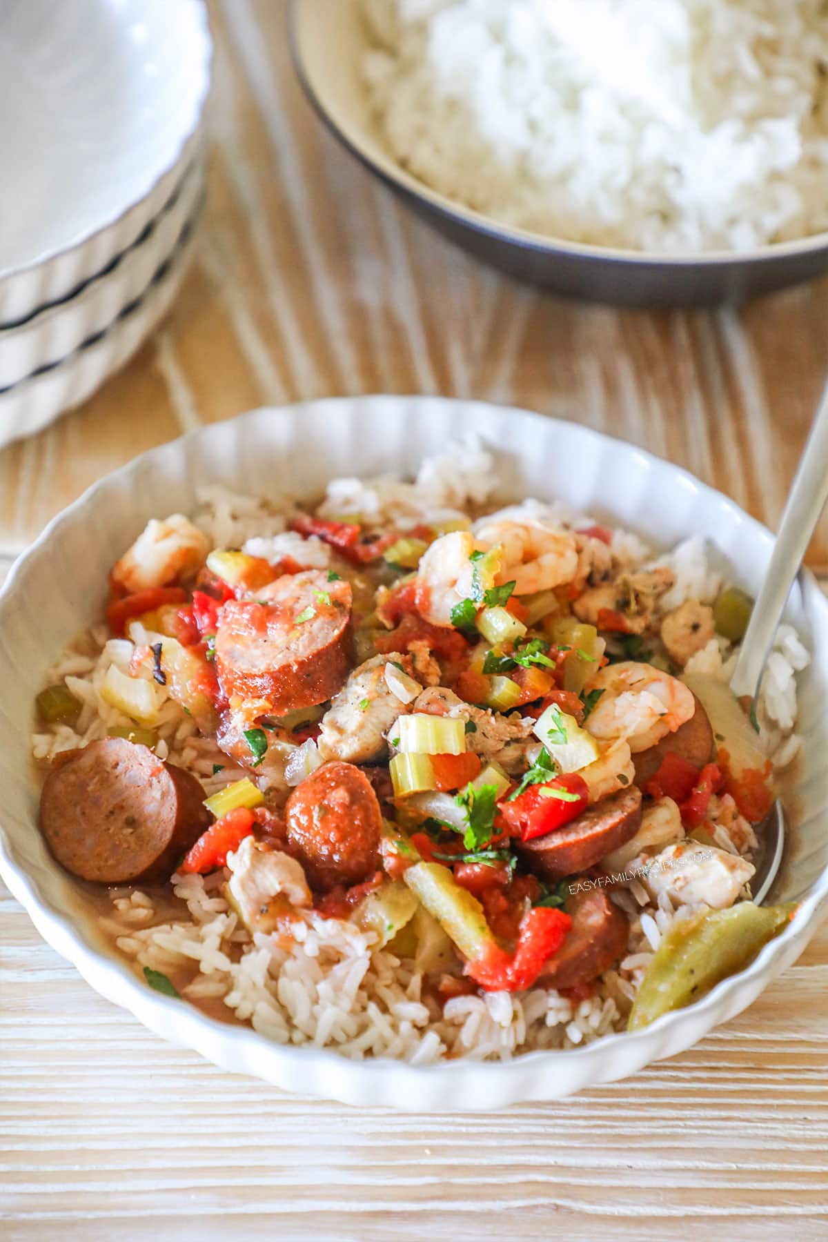 Jambalaya served over rice in a white bowl. On a wooden background with a stack of white plates and bowl of rice in the background.