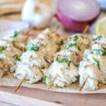 greek chicken gyro skewers on wooden board- red onion, garlic and pita bread in background