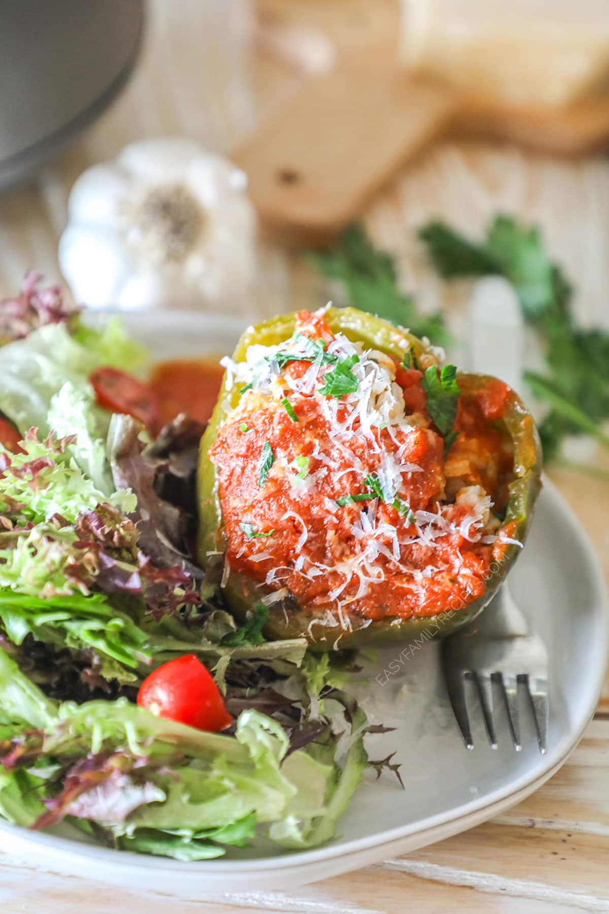 Stuffed pepper on a white plate with side salad and a fork. On a wooden background with garlic head, fresh parsley and wooden board.
