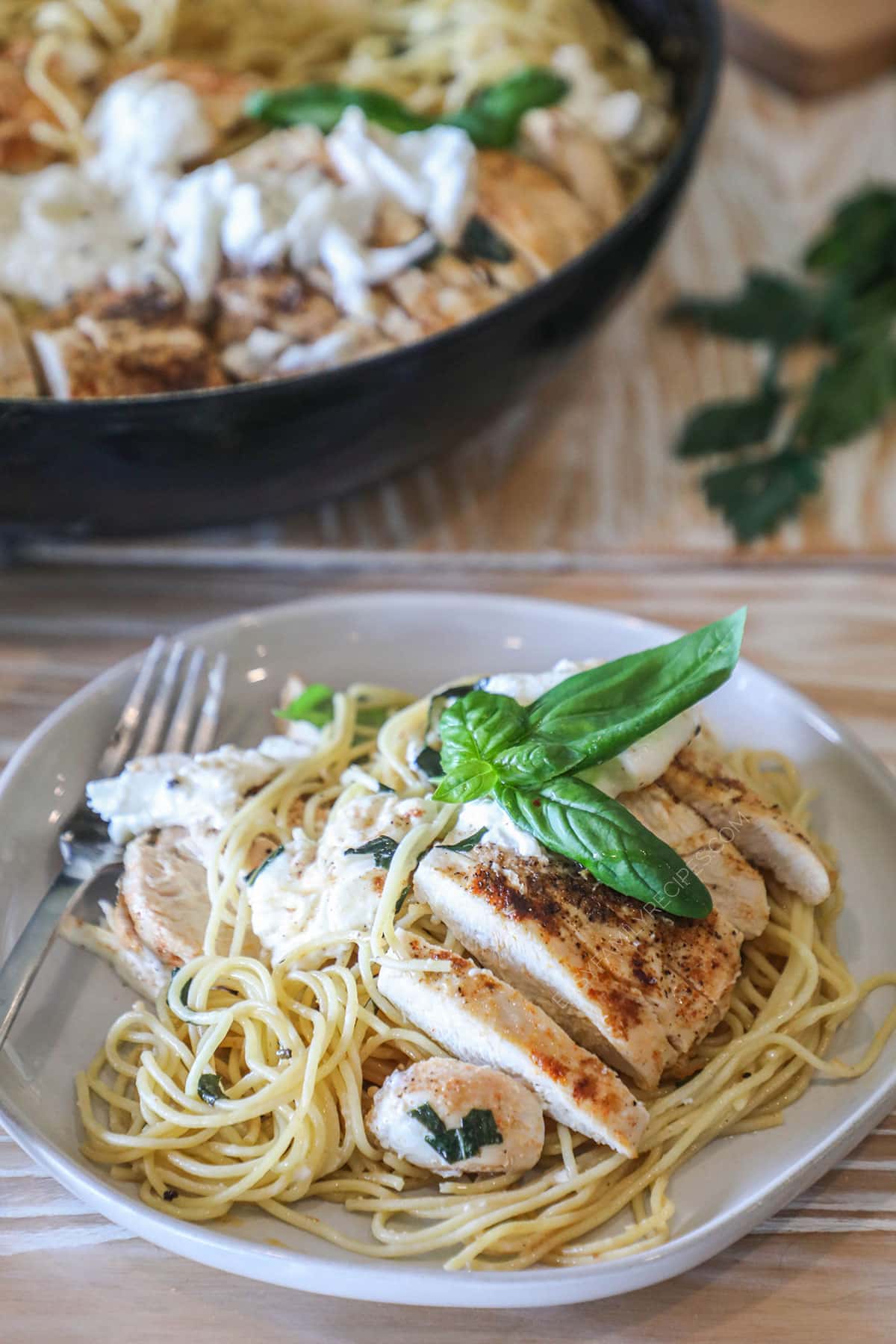 Serving of brown butter spaghetti with sliced chicken, burrata and basil on top. On a white dish with a fork on the side. Full pan of spaghetti meal in background. On a wooden table