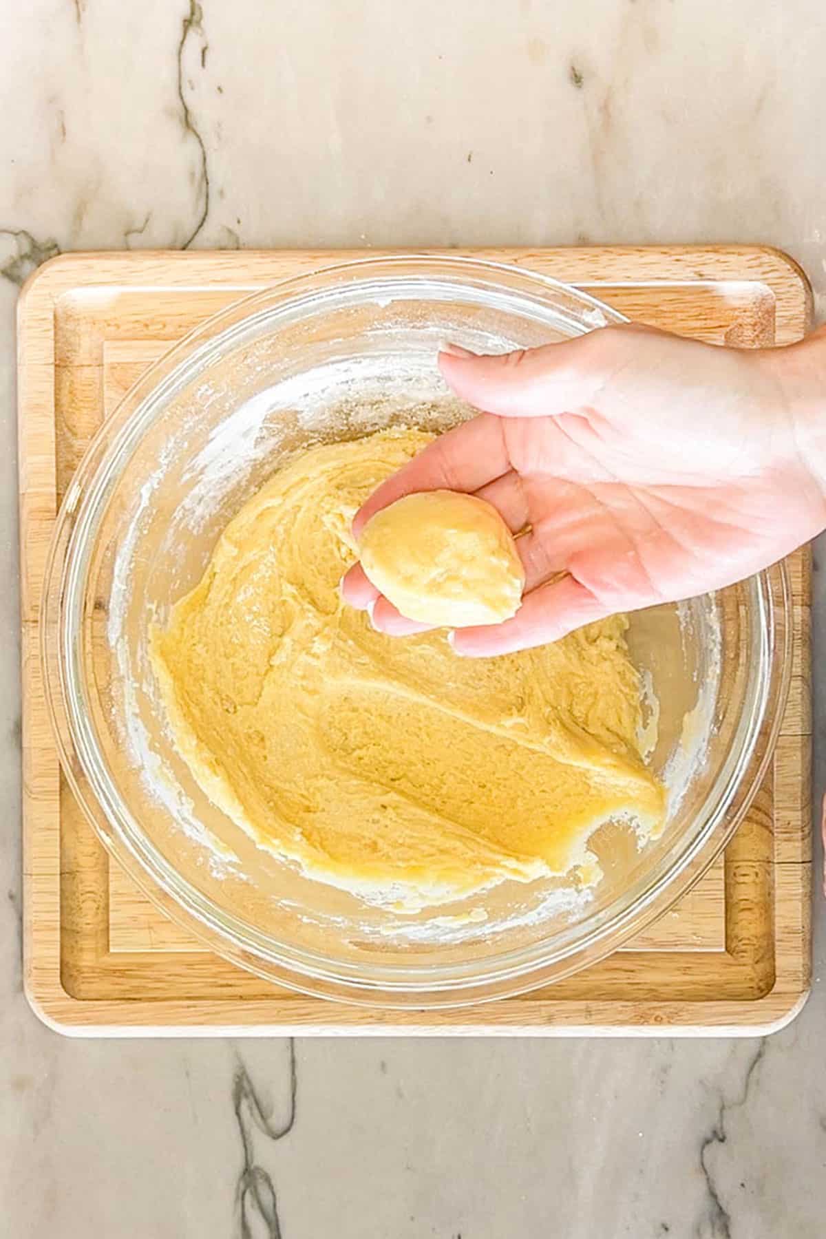 Lemon cake mix cookie dough in a glass bowl. Hand holding formed dough ball. On a wooden board and background.