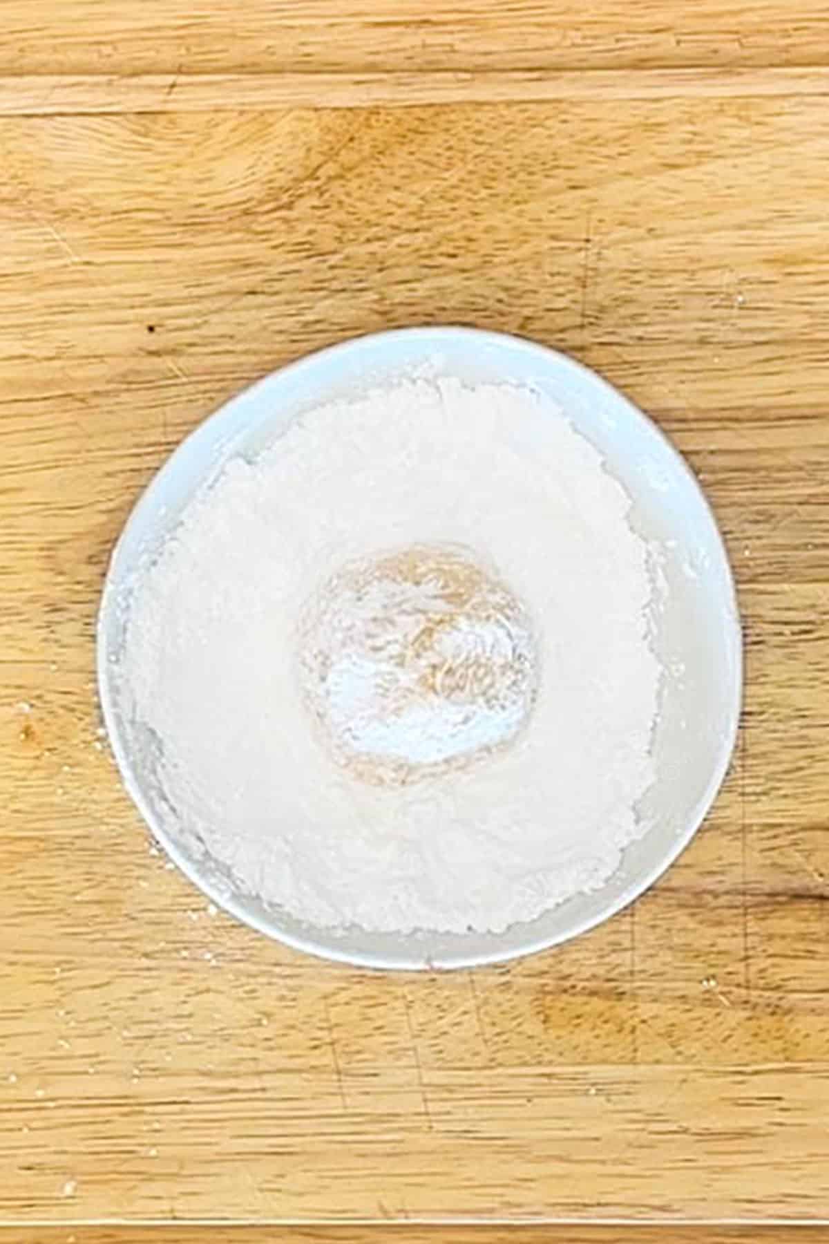 White bowl with powdered sugar. Cookie dough ball in middle. On a wooden background