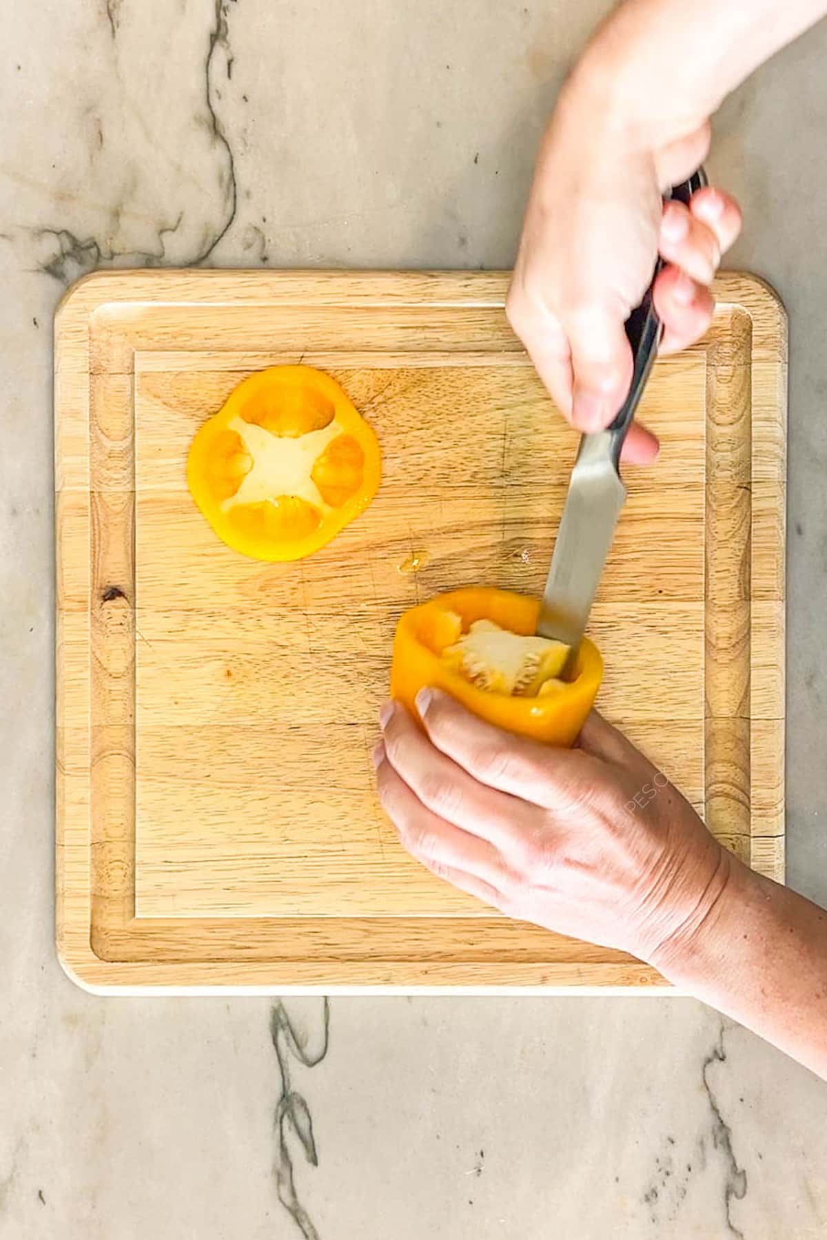 Process picture of hollowing out a bell pepper. Two hands are holding and using a knife to hollow out pepper. On a wooden board with a marble background.