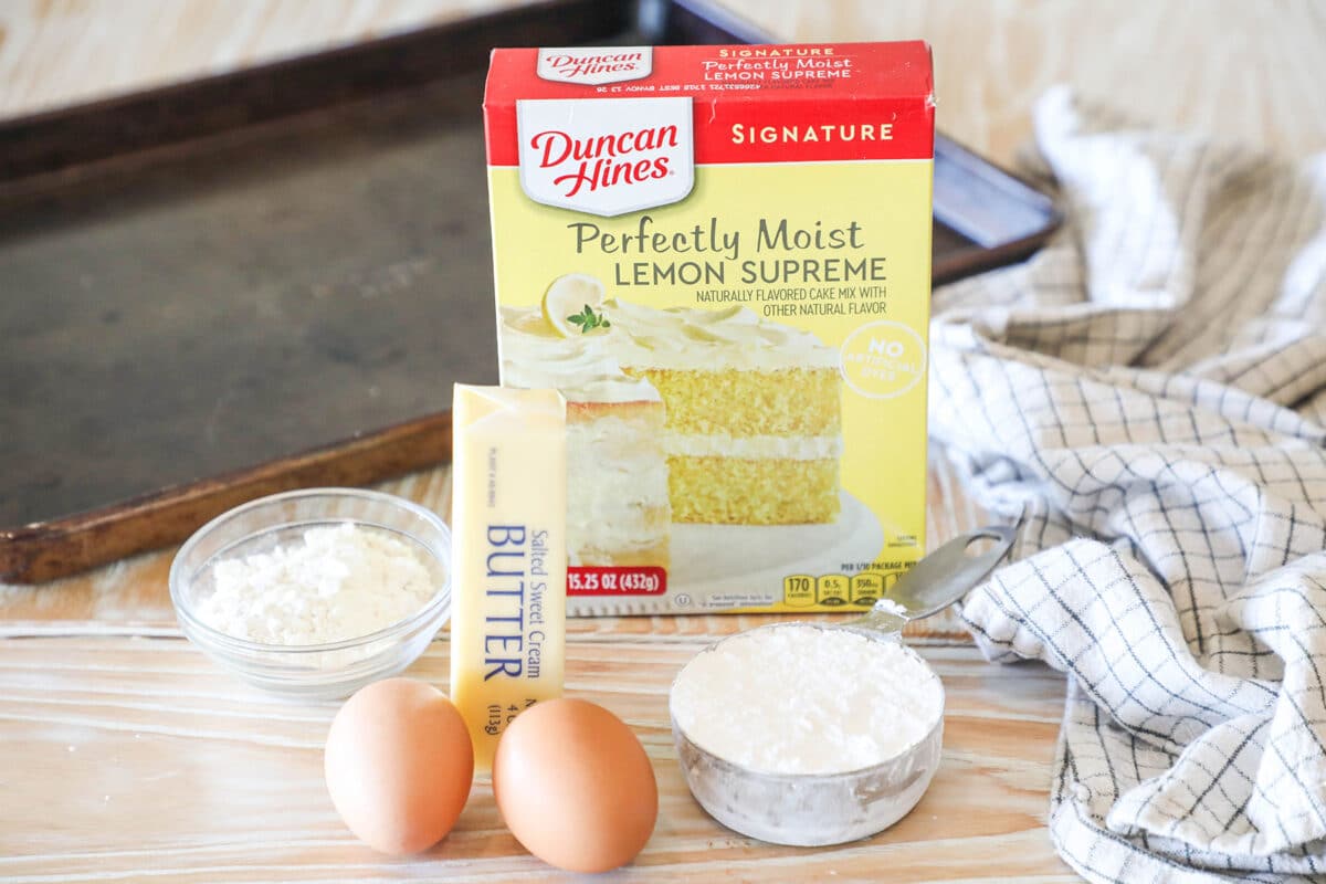 Ingredients for lemon cake mix crinkle cookies. Box of cake mix, two eggs, stick of butter, bowl of flour, bowl of powdered sugar. On a wooden table with a sheet pan and kitchen towel.