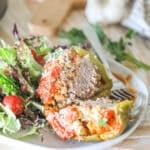 Cooked stuffed pepper, cut in half. On white plate with side salad. On a wooden table with garlic and wooden board in background.