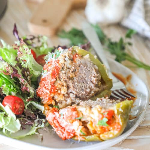 Cooked stuffed pepper, cut in half. On white plate with side salad. On a wooden table with garlic and wooden board in background.