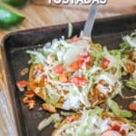 a chicken tostada topped with lettuce and tomato being lifted off of a baking sheet.