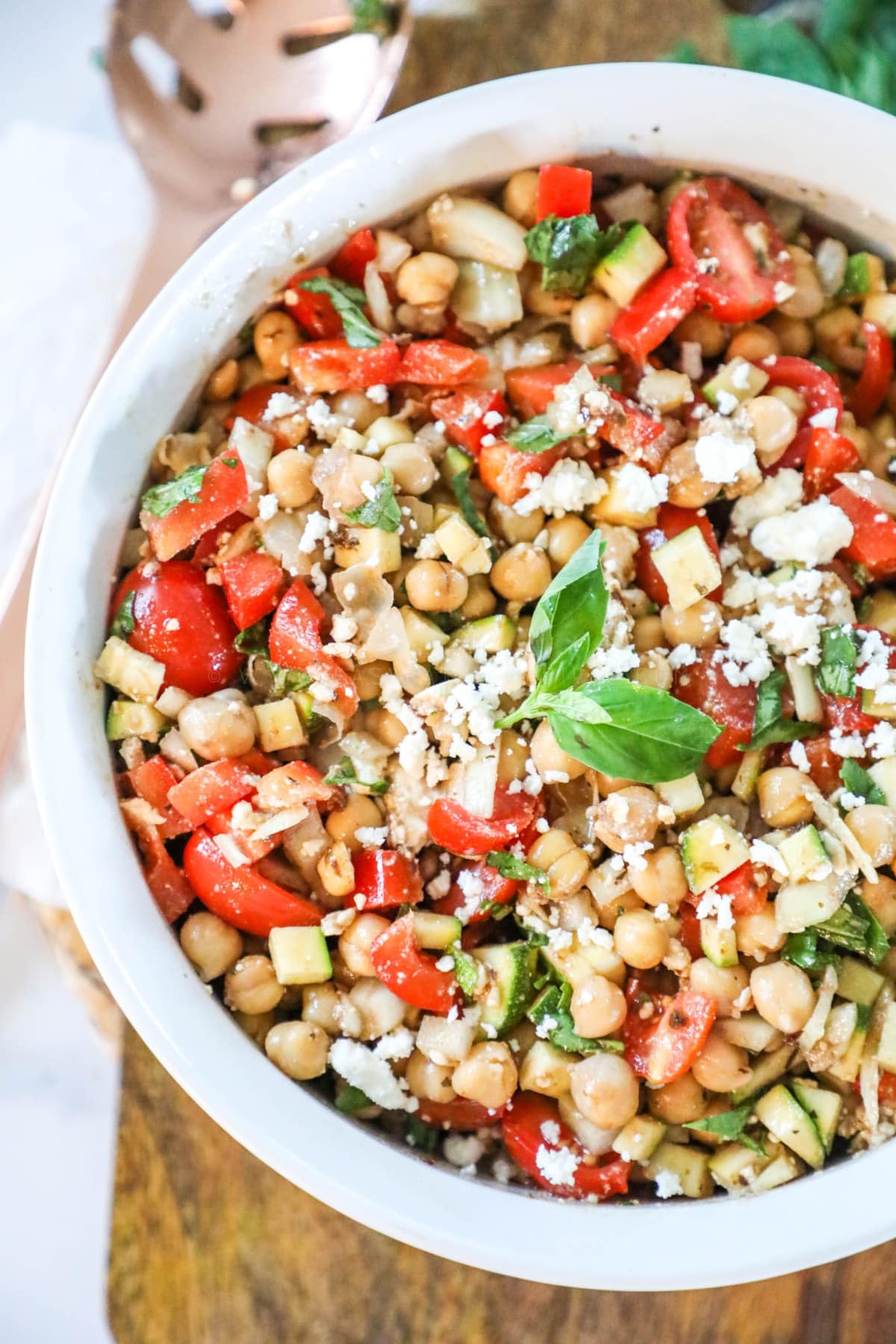 a white ceramic bowl of mediterranean chickpea salad, sitting on a wooden cutting board