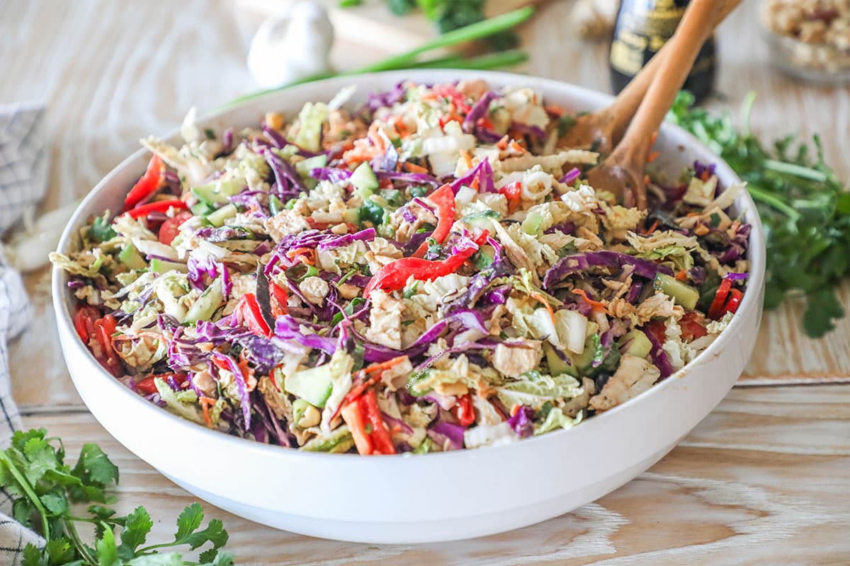 Thai Style Chicken Salad with Peanut Dressing in a large white bowl with wooden spoons. On a wooden background with fresh herbs