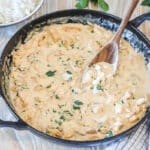 Smothered Chicken in pan with wooden spoon. On a wooden background with kitchen towel, fresh herbs and bowl of rice