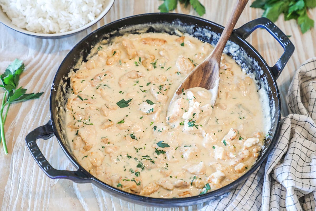 Smothered Chicken in pan with wooden spoon. On a wooden background with kitchen towel, fresh herbs and bowl of rice