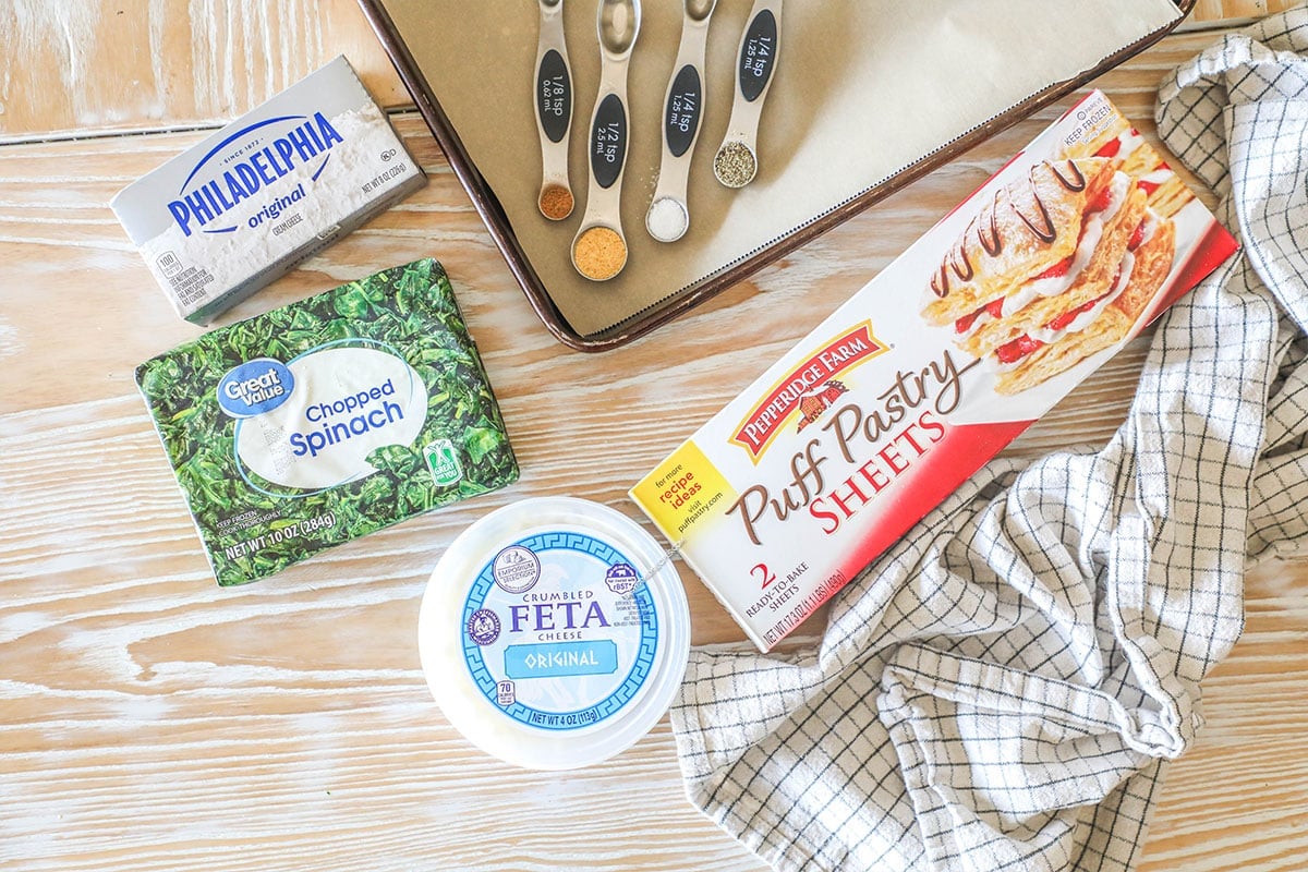 Ingredients needed to make Spinach feta puff pastry swirls. Spinach, feta, cream cheese, seasonings and puff pastry. On a wooden background with a kitchen towel.