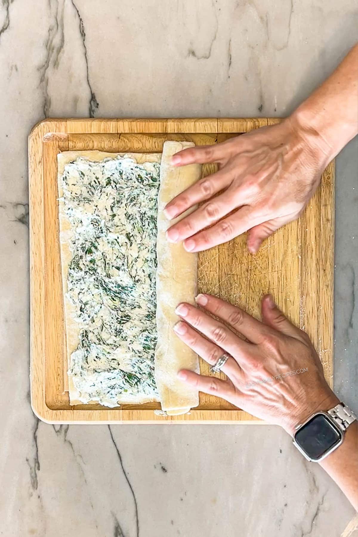 Pastry being rolled by two hands into a log. On a wooden board.