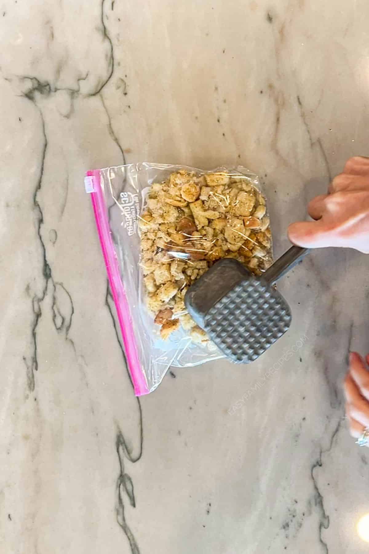 Step one of french onion chicken bake- bag of croutons being crushed by hammer- on marble background