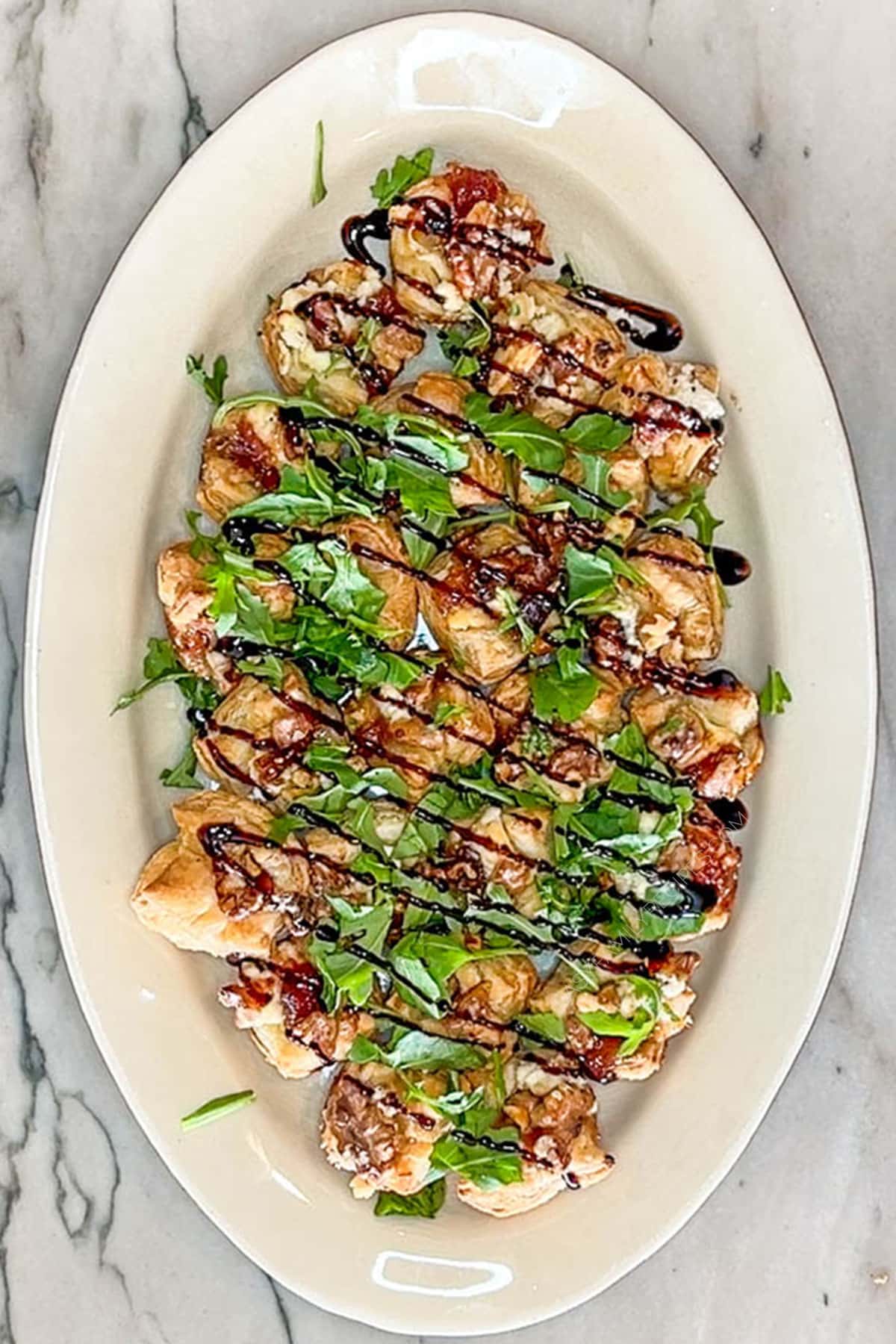 Goat Cheese and Fig Jam Puff Pastry Bites with balsamic glaze drizzled on top. On a white dish and a marble background