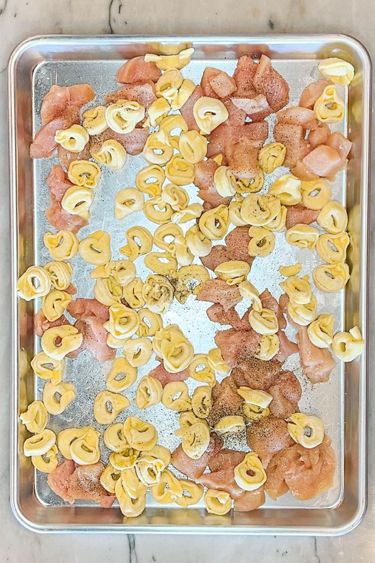 Tortellini and seasoned diced chicken breast placed on a sheet pan to cook.