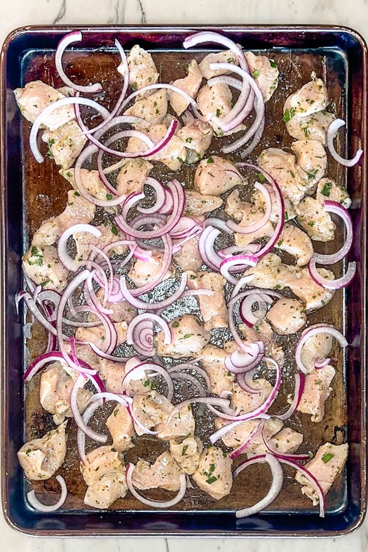 Pieces of marinated chicken breast and slices red onions placed on the sheet pan.