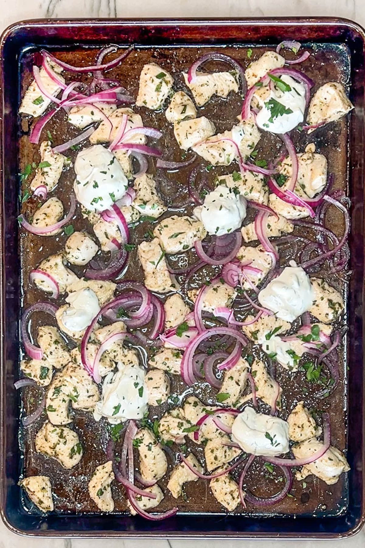Baked Mediterranean chicken with red onions on a sheet pan, topped with dollops of tzatziki.